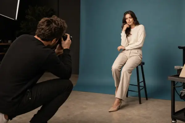 Gutscheine 2 Professionelles Portrait-Fotoshooting bei PM STUDIOS München: Ein Fotograf fotografiert eine sitzende Frau in hellem Outfit vor einem modernen blau-grauen Studiohintergrund; authentische Einblicke in die Studioarbeit.